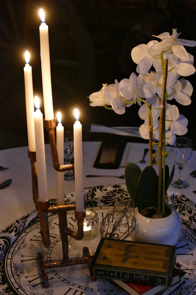 Close-up of a dinner table centerpiece featuring lit white candles in a copper pipe holder, a white orchid in a ceramic pot, and vintage books set on a clock-face base.