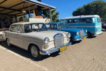 Three vintage vehicles, including a beige Mercedes-Benz Fintail, a blue Mercedes-Benz Ponton, and a light blue Volkswagen Type 2 bus, parked on a brick driveway under a clear blue sky.