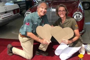 A man and a woman smiling and kneeling on a red carpet while holding large cardboard hearts in front of a classic red Mercedes-Benz.