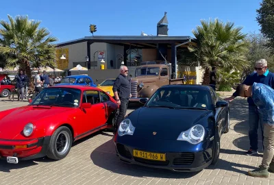 A classic red Porsche 911 and a dark blue Porsche 996 parked side-by-side at the Old Wheelers Club, with a vintage brown pickup truck and palm trees in the background.