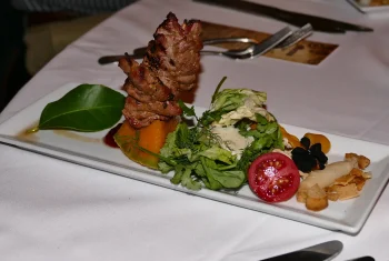 A gourmet appetizer plated on a white rectangular dish featuring a grilled meat skewer on a roasted pumpkin wedge, served with a fresh green salad and a halved cherry tomato.
