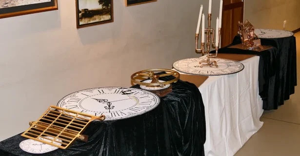 A display table at an event featuring industrial-style decor, including copper pipe candelabras, vintage car parts, and large clock-face placemats against a gallery wall of historic photos.
