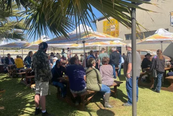 People gathered at wooden picnic tables under yellow and white umbrellas in a sunlit outdoor garden area at the Old Wheelers Club.