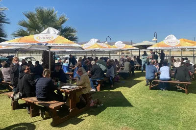 A crowded outdoor beer garden with people sitting at wooden picnic tables at the Old Wheelers Club on a sunny day.