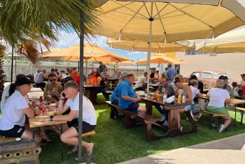 Crowded outdoor restaurant patio with picnic tables, sun umbrellas, and a vintage car backdrop in Namibia.