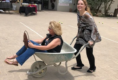 Two women laughing during a playful wheelbarrow race at the Old Wheelers Club in Windhoek, Namibia, with one woman "steering" using a vintage metal wheel while being pushed.