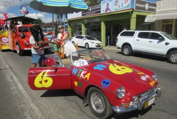 A red MG MGB convertible decorated with colorful "WIKA" stickers and emojis participating in a street parade, with a person in costume standing beside it under a striped umbrella.